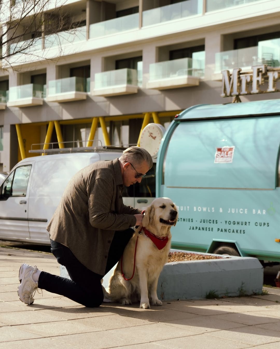 Street photo by Chiemeka Madufor: a man in sunglasses kneels to pet a golden retriever in a red bandana beside a juice bar van