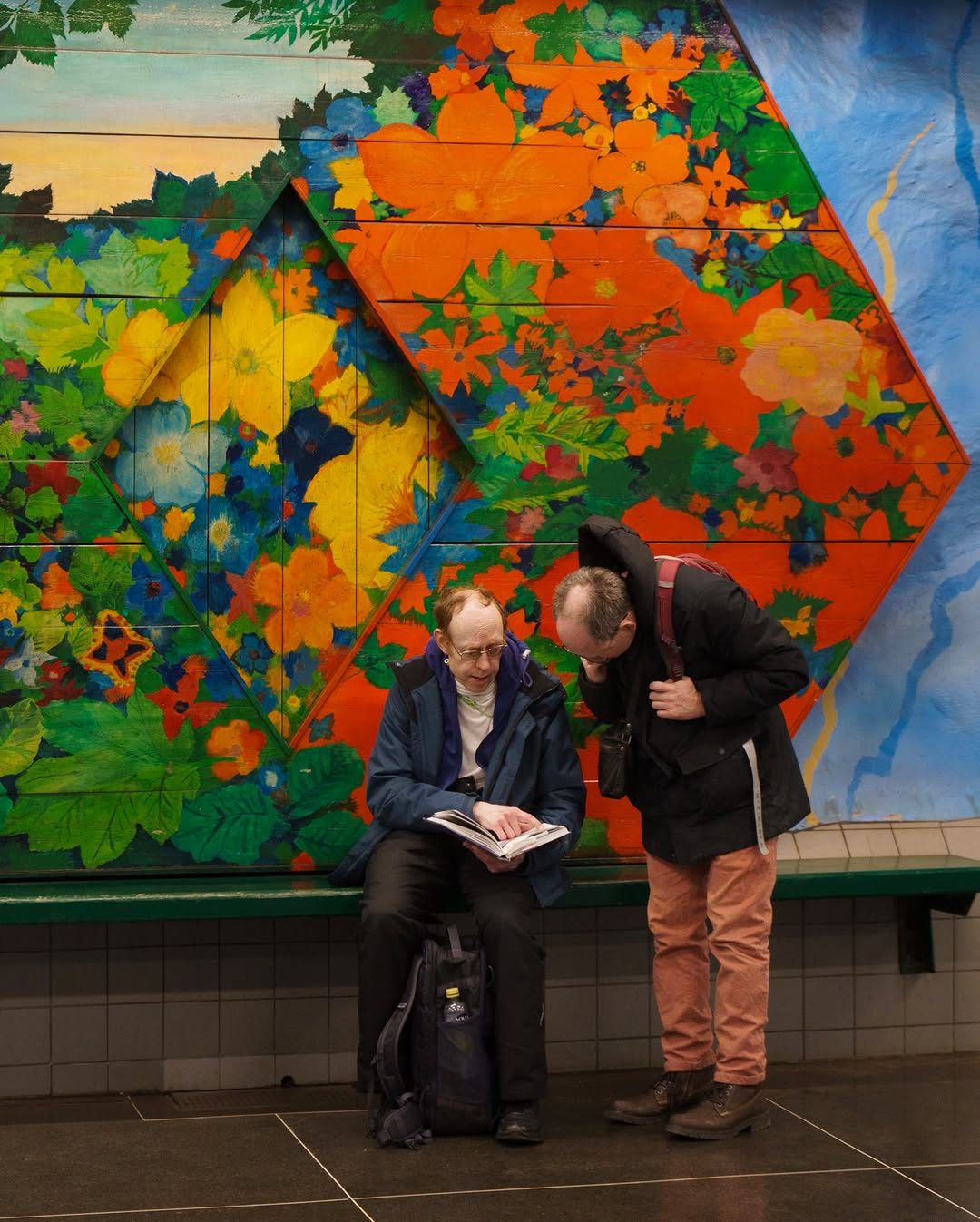 Street photo by Chiemeka Madufor: two older men read a book together on a bench in front of a bright floral mural